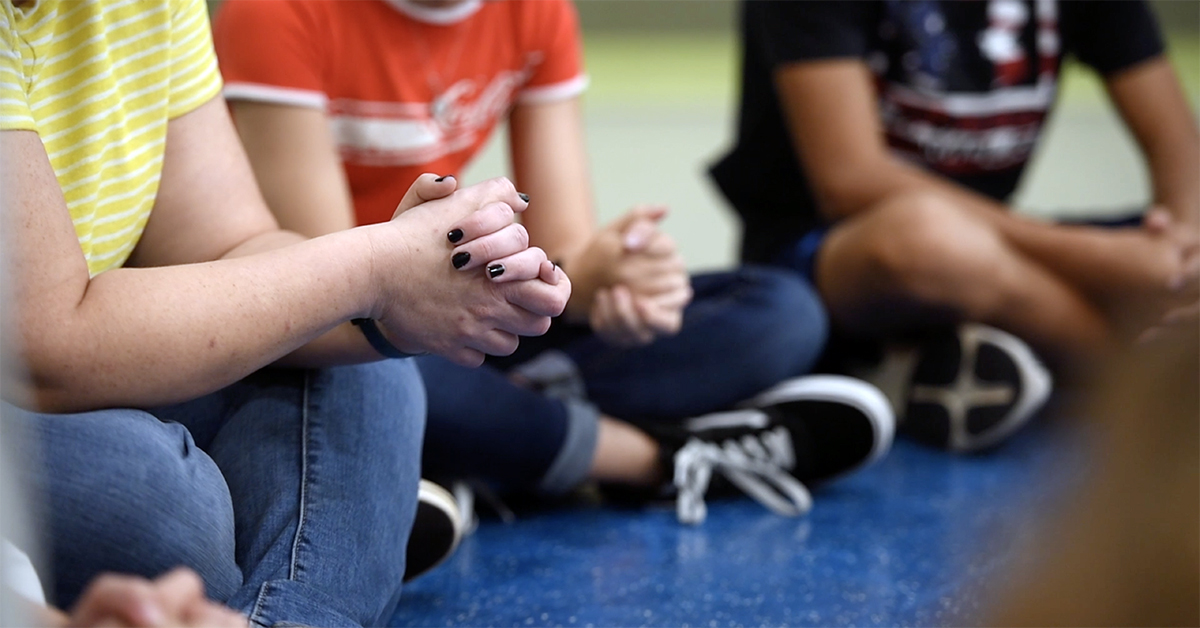 Photo of an adult and a children sitting cross-legged, with hands folded in prayer