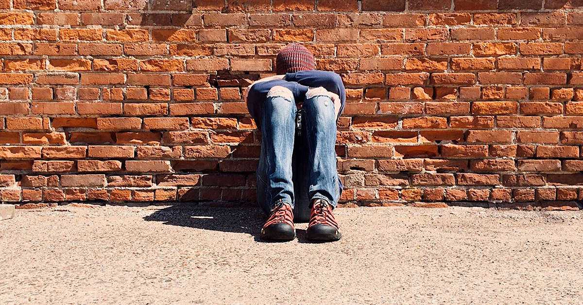 Photo of a teen sitting against a brick wall with their head against their folded arms.